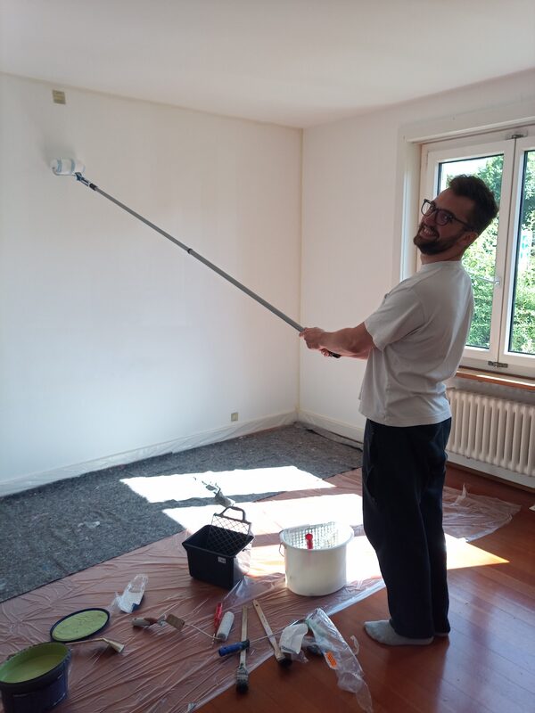 Syd, holding a paint roller against a wall and grinning at the camera